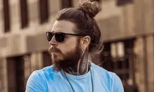 Hombre con pelo largo recogido en un moño, barba, gafas de sol y tatuajes en el cuello.