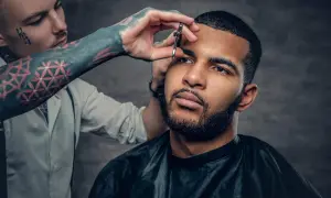 Barbero con tatuajes en el brazo recorta el **buzz cut corto** de un hombre con barba.