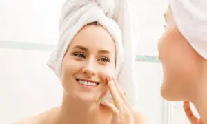 Mujer sonriente con toalla en la cabeza aplicándose crema en la cara. Una rutina de cara para piel grasa que deja la piel radiante.