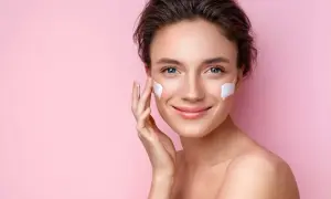 Mujer sonriente aplicándose crema en las mejillas, esencial para los cuidados de la piel en verano.