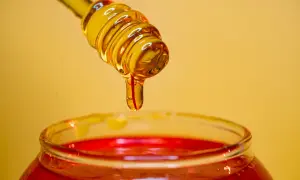 Miel dorada goteando de un agitador sobre un frasco rojo, ideal para un exfoliante facial casero.