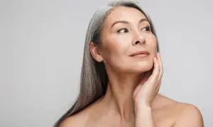 Mujer asiática con cabello gris plateado, tocando su rostro con la mano. Su mirada se dirige hacia la derecha, mostrando una piel radiante y un ejemplo de well aging.