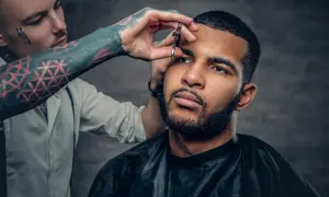 Barbero con tatuajes en el brazo recorta las cejas a un hombre con un corte buzz cut y barba.
