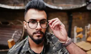 Hombre con barba y gafas, luciendo un moderno corte de pelo mohicano y chaqueta de camuflaje.