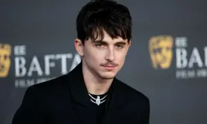 Joven con un corte de pelo hombre medio largo, peinado hacia adelante con flequillo, luciendo un traje negro y un collar brillante.