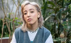 Joven con un moderno corte de pelo mullet mujer, luce un collar plateado y un chaleco azul sobre una camiseta blanca, rodeada de vegetación.