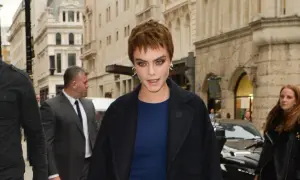 Cara Delevingne con un atrevido buzz cut largo, mirada intensa y abrigo oscuro, rodeada de seguridad en una calle urbana.