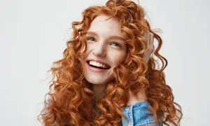 Mujer joven con cabello rojo y rizado, sonriendo y tocándose el pelo. Su melena merodo curly es espectacular.