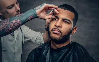 Barbero con tatuajes en el brazo recorta las cejas a un hombre con un corte buzz cut y barba.
