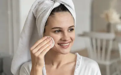 Mujer sonriente con toalla en la cabeza, aplicándose un disco de algodón en la mejilla. Un ejemplo de limpieza facial antes y después.