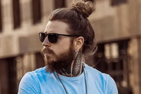 Hombre con pelo largo recogido en un moño, barba, gafas de sol y tatuajes en el cuello.