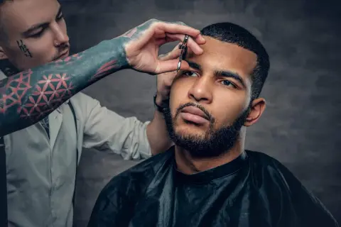 Barbero con tatuajes en el brazo recorta el **buzz cut corto** de un hombre con barba.