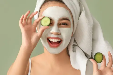 Mujer sonriente con mascarilla facial y rodajas de pepino, disfrutando de tratamientos faciales naturales.
