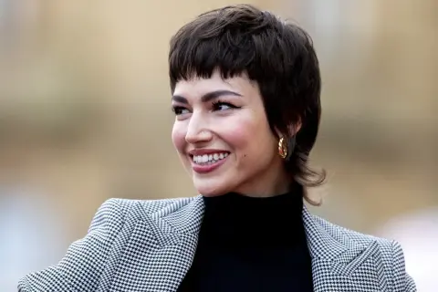 Mujer sonríe con un corte pixie medio, luciendo un blazer de pata de gallo y un pendiente dorado.
