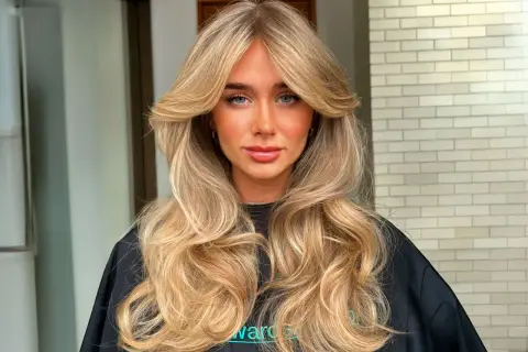 Mujer con cabello rubio largo y ondulado, con un flequillo mariposa que enmarca su rostro.
