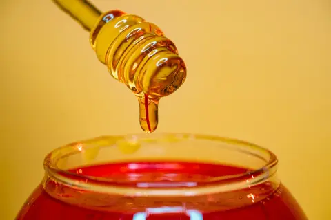 Miel dorada goteando de un agitador sobre un frasco rojo, ideal para un exfoliante facial casero.