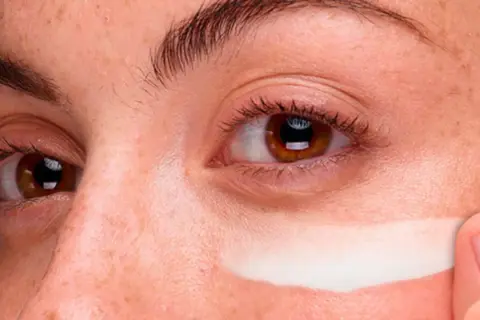 Dedo aplica crema contorno de ojos en toda la cara, cubriendo pecas y piel.
