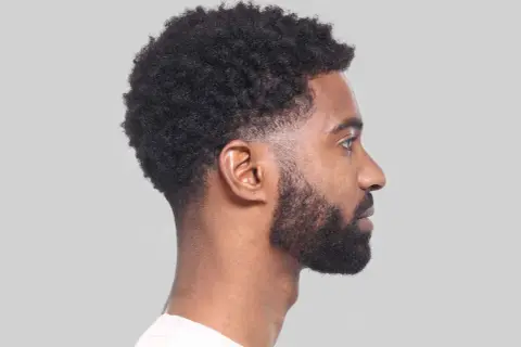 Perfil de hombre con cabello rizado y barba, luciendo un moderno corte taper fade.