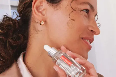 Mujer con cabello rizado y aretes dorados sostiene un frasco de sérum, mostrando cómo hidratar la piel desde dentro.