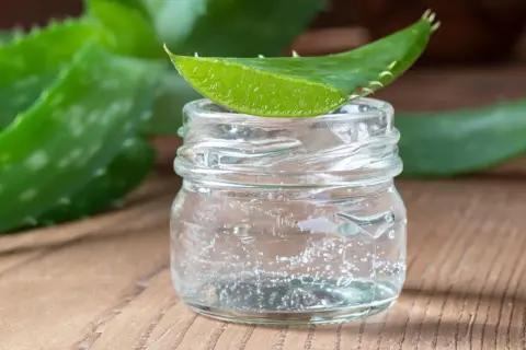 Un trozo de hoja de aloe vera descansa sobre un frasco de vidrio con gel transparente, ideal para un tratamiento de aloe vera para el cabello sin enjuagar.