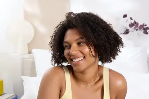Mujer sonriente con cabello rizado y una permanente ondulada, luciendo un top amarillo en una habitación luminosa.