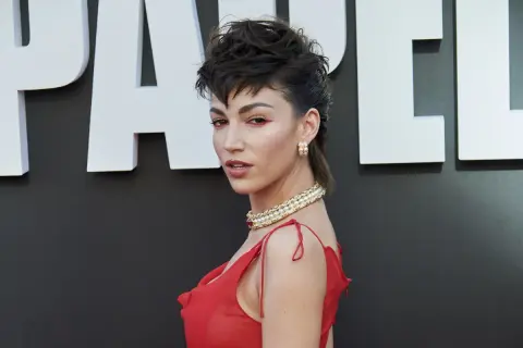 Mujer con un atrevido corte mullet y maquillaje dramático, luciendo un vestido rojo y un collar de perlas.