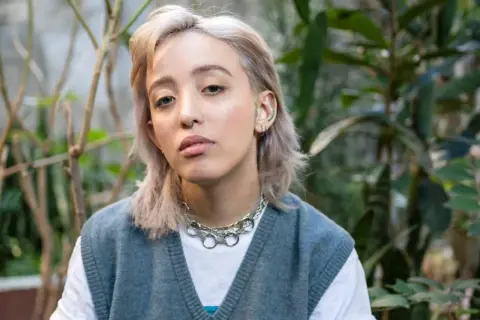 Joven con un moderno corte de pelo mullet mujer, luce un collar plateado y un chaleco azul sobre una camiseta blanca, rodeada de vegetación.