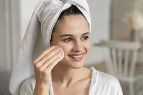 Mujer sonriente con toalla en la cabeza, aplicándose un disco de algodón en la mejilla. Un ejemplo de limpieza facial antes y después.