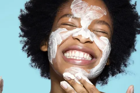 Una mujer sonríe con una mascarilla blanca en la cara. ¿Qué va primero el exfoliante o el limpiador?
