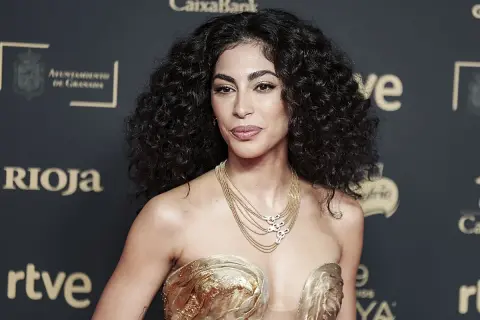 Mujer con cabello oscuro y rizos definidos, luciendo un vestido dorado y un collar de cadenas.