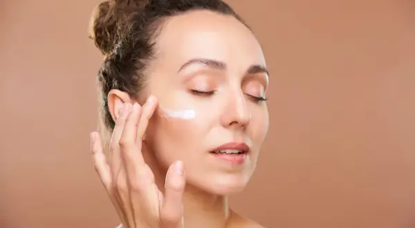 Mujer aplicándose crema en la mejilla, cuidando su piel con retinol en verano.