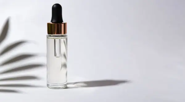 Frasco de vidrio con gotero, contiene un suero transparente, posiblemente con ácido ferúlico, sobre fondo blanco con sombras de hojas.
