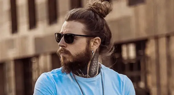 Hombre con pelo largo recogido en un moño, barba, gafas de sol y tatuajes en el cuello.