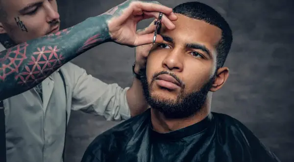 Barbero con tatuajes en el brazo recorta el **buzz cut corto** de un hombre con barba.