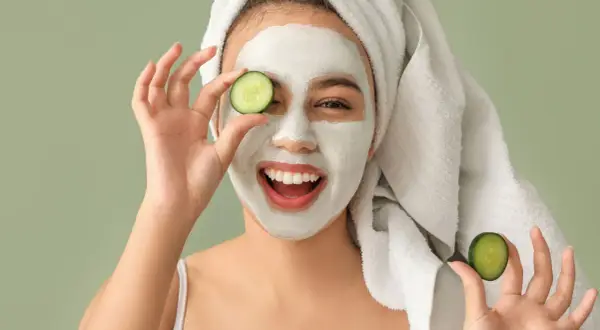 Mujer sonriente con mascarilla facial y rodajas de pepino, disfrutando de tratamientos faciales naturales.