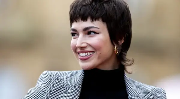 Mujer sonríe con un corte pixie medio, luciendo un blazer de pata de gallo y un pendiente dorado.