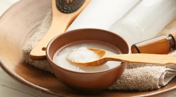 Un tazón con agua de arroz para la cara, una cuchara de madera con arroz, un cepillo para el cabello y botellas de productos de belleza.