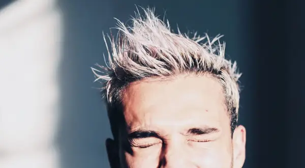 Hombre con pelo rubio despeinado y ojos cerrados. Consejos sobre como hidratar el pelo hombre para un look fresco.