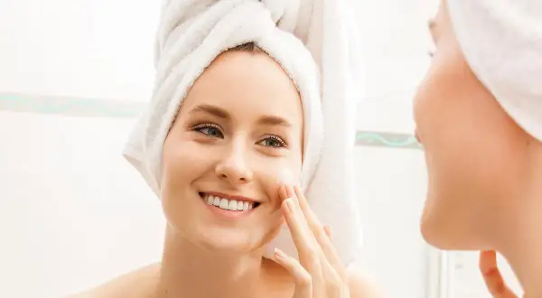 Mujer sonriente con toalla en la cabeza aplicándose crema en la cara. Una rutina de cara para piel grasa que deja la piel radiante.