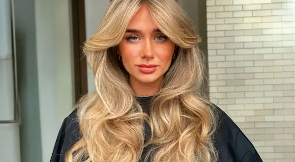 Mujer con cabello rubio largo y ondulado, con un flequillo mariposa que enmarca su rostro.