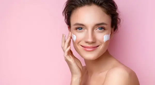 Mujer sonriente aplicándose crema en las mejillas, esencial para los cuidados de la piel en verano.