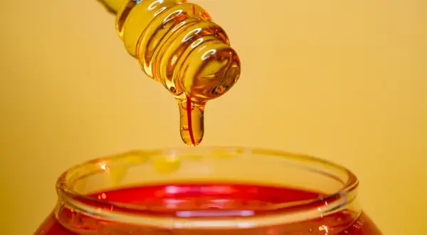 Miel dorada goteando de un agitador sobre un frasco rojo, ideal para un exfoliante facial casero.