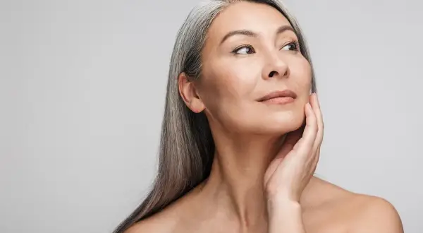 Mujer asiática con cabello gris plateado, tocando su rostro con la mano. Su mirada se dirige hacia la derecha, mostrando una piel radiante y un ejemplo de well aging.