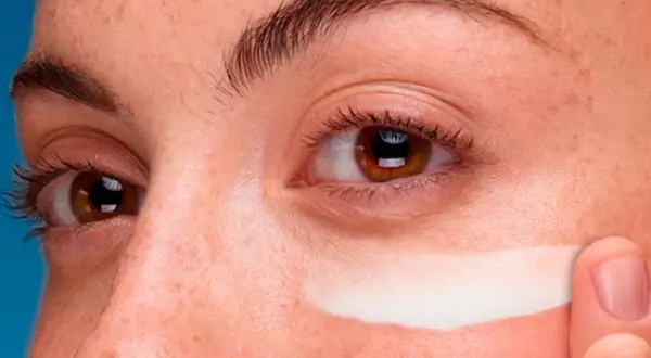 Dedo aplica crema contorno de ojos en toda la cara, cubriendo pecas y piel.