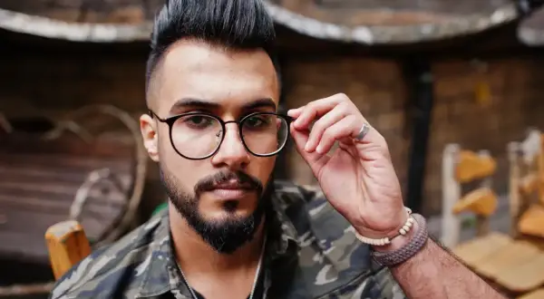 Hombre con barba y gafas, luciendo un moderno corte de pelo mohicano y chaqueta de camuflaje.