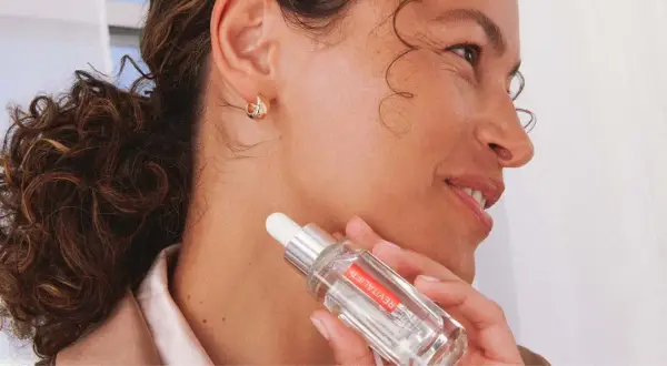 Mujer con cabello rizado y aretes dorados sostiene un frasco de sérum, mostrando cómo hidratar la piel desde dentro.