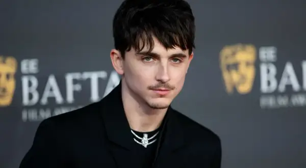 Joven con un corte de pelo hombre medio largo, peinado hacia adelante con flequillo, luciendo un traje negro y un collar brillante.