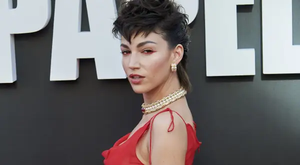 Mujer con un atrevido corte mullet y maquillaje dramático, luciendo un vestido rojo y un collar de perlas.