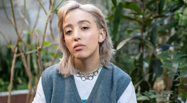 Joven con un moderno corte de pelo mullet mujer, luce un collar plateado y un chaleco azul sobre una camiseta blanca, rodeada de vegetación.