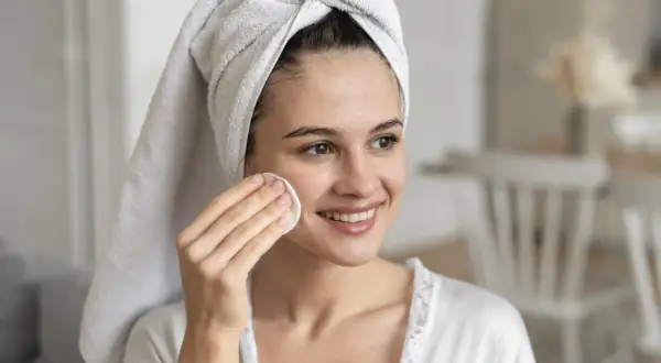 Mujer sonriente con toalla en la cabeza, aplicándose un disco de algodón en la mejilla. Un ejemplo de limpieza facial antes y después.
